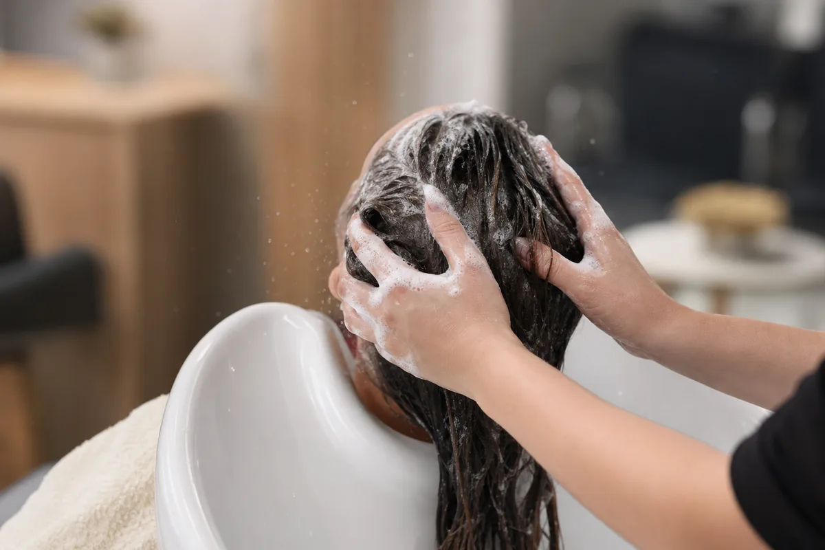 Vue de dos d’une cliente installée au bac de lavage d’un salon, mousse et jets d’eau, lumière douce