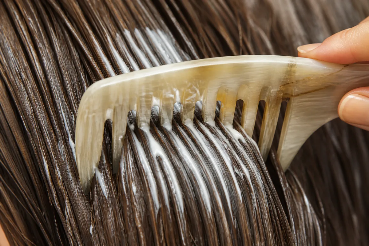 Peigne à dents larges répartissant un masque blanc sur des longueurs de cheveux bruns humides